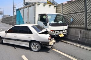 A white sedan with severe rear-end damage is shown after being struck by a delivery truck on a highway, both vehicles up against a concrete barrier.